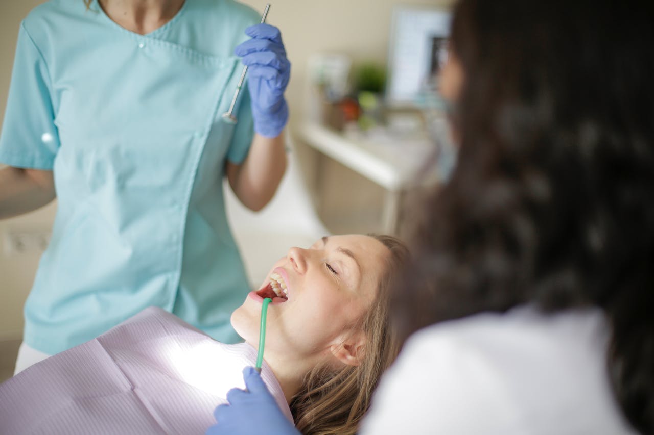 Crop faceless female dentist in uniform holding mouth mirror while preparing for inspecting female patient teeth with assistant holding tube suction in patient mouth